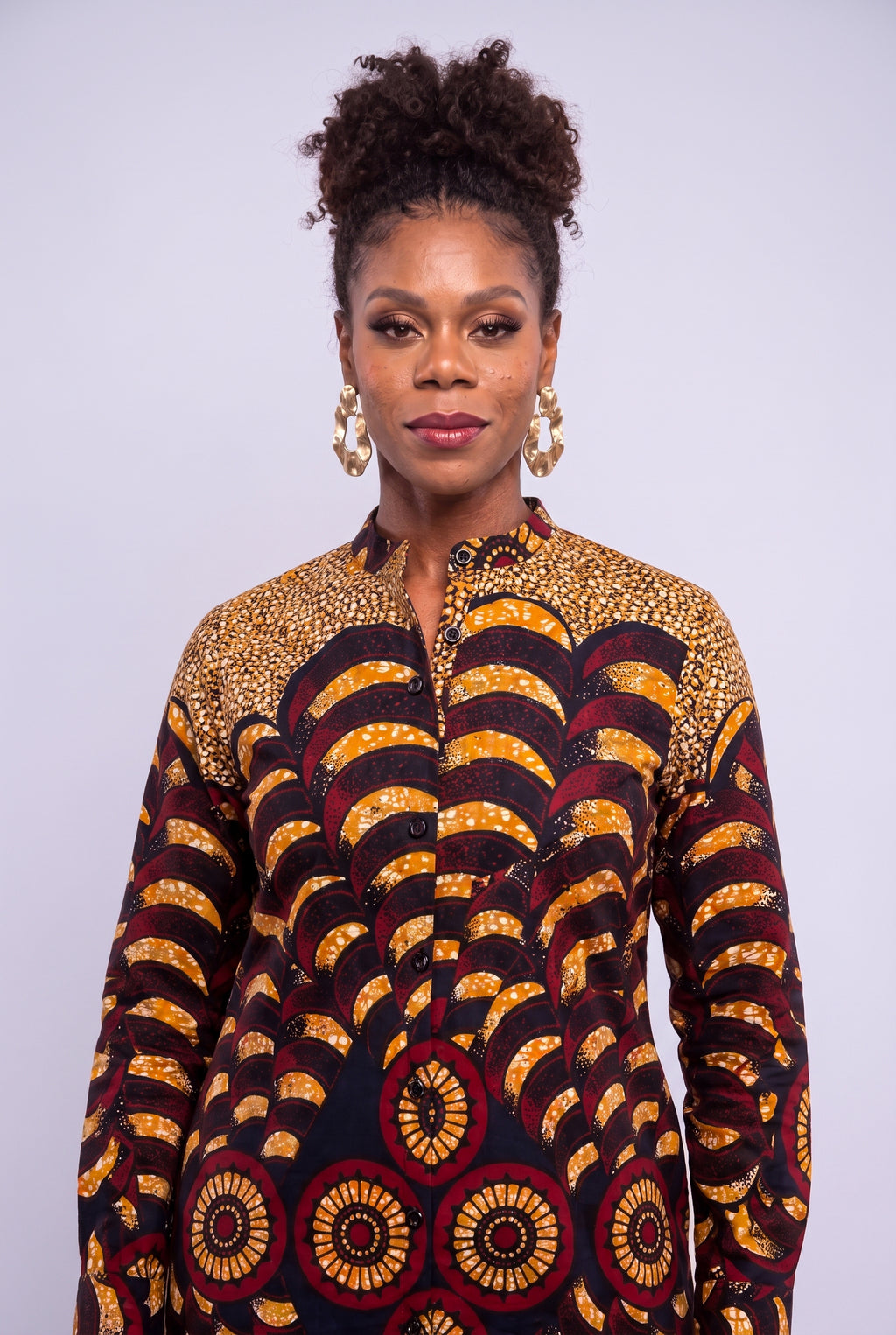 A woman with her hair in a high bun wears a long-sleeve button-up shirt featuring a bold African wax print pattern with yellow, red, and black scalloped waves and circular motifs, set against a plain light gray studio background.