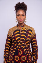 A woman with her hair in a high bun wears a long-sleeve button-up shirt featuring a bold African wax print pattern with yellow, red, and black scalloped waves and circular motifs, set against a plain light gray studio background.