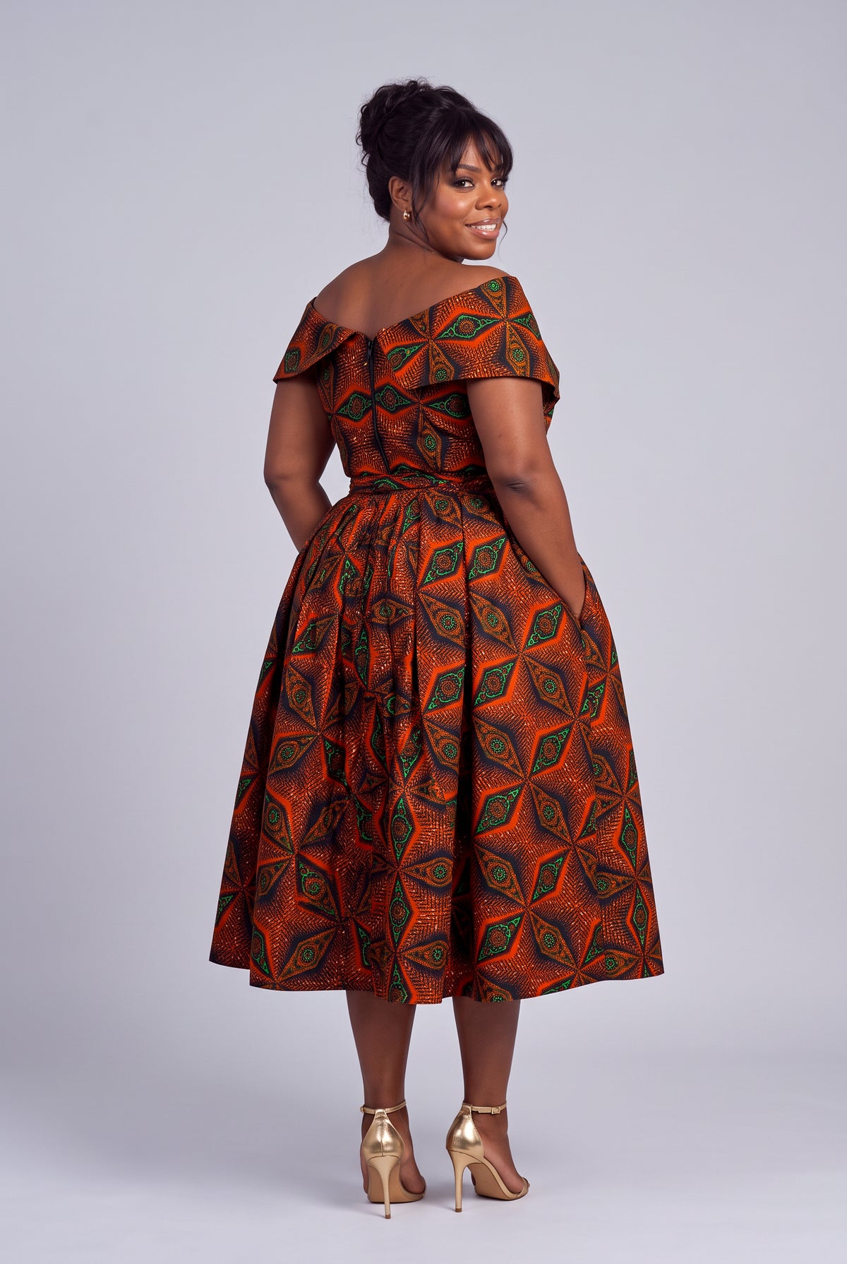 Back view of  woman modeling an off-the-shoulder Ankara midi dress with a vibrant orange, brown, and green geometric pattern