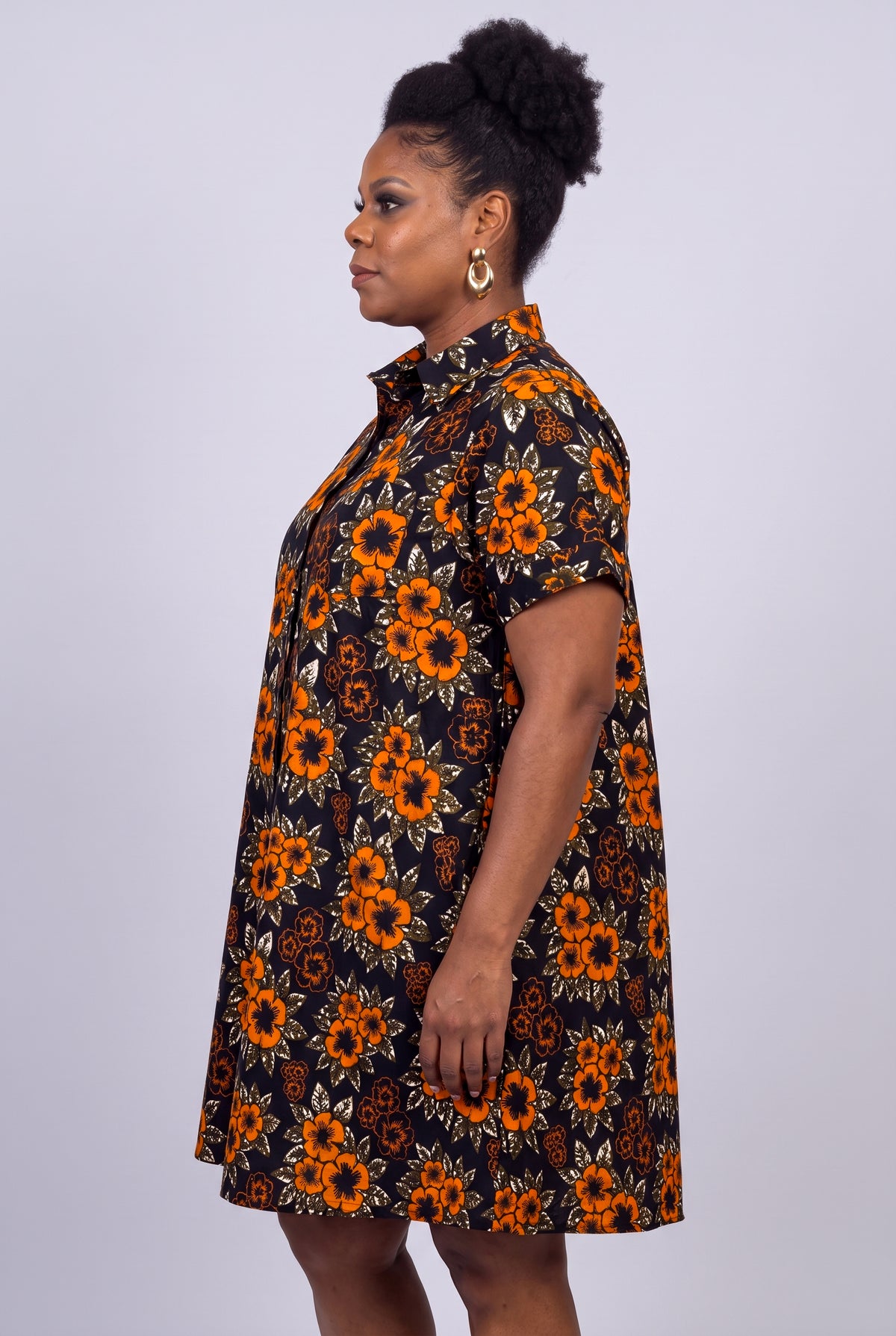A woman shown in side profile wearing a short-sleeve button-up African shirt dress with an orange and yellow floral print on a black background,