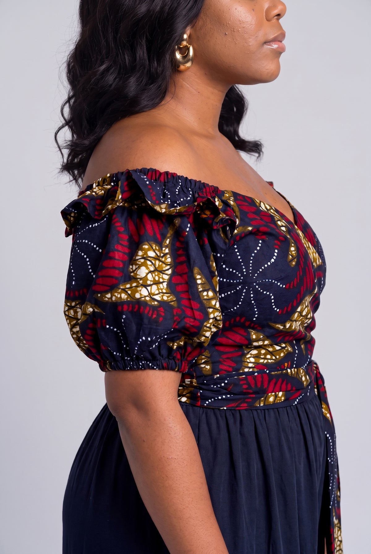 Side profile view of a curvy woman wearing a red, gold and black African print off-the-shoulder wrap top with navy blue trousers