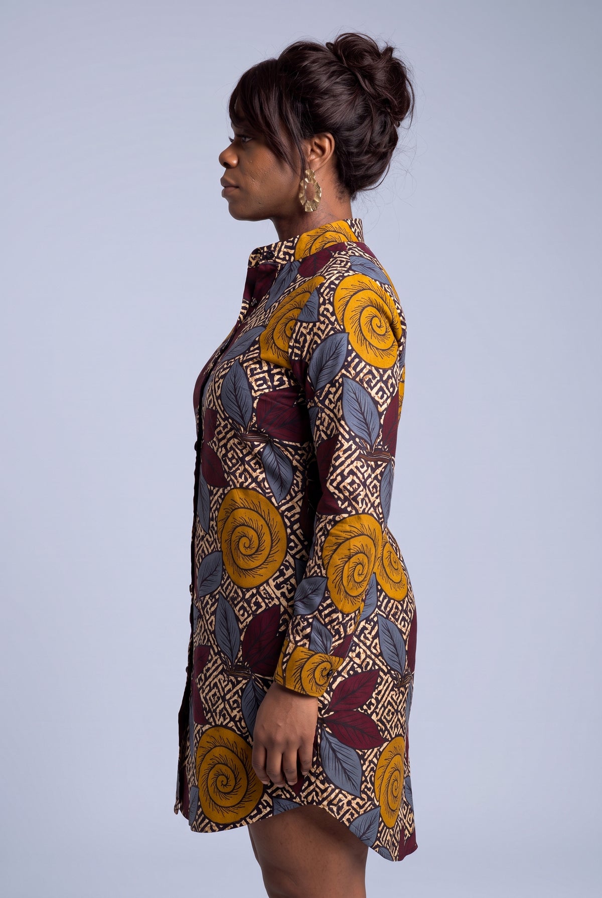 A woman shown in side profile wearing a long-sleeve button-front shirt dress with an African wax print pattern featuring yellow swirls, burgundy leaves, and beige geometric borders on a light gray studio background.