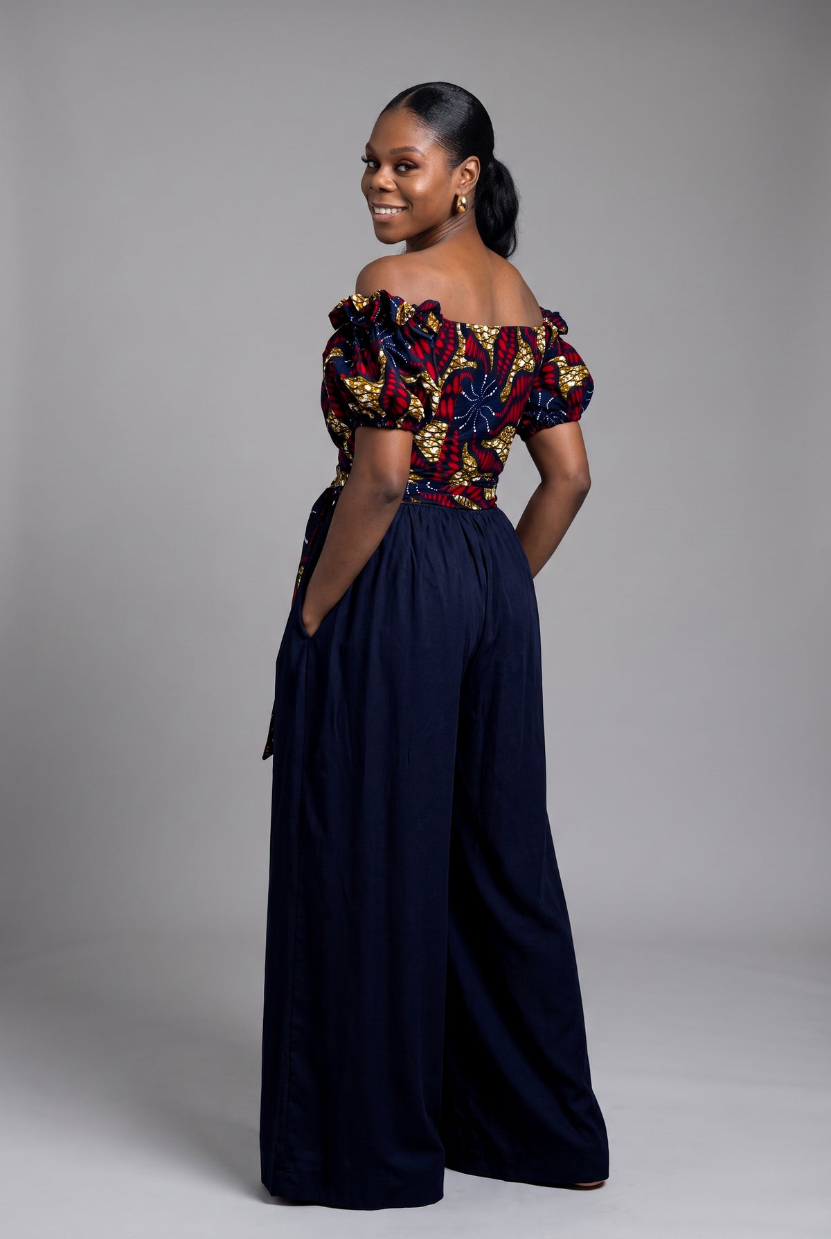 Back view of a tailored off shoulder wrap Ankara top in vibrant red and navy blue wax print and Navy Trouser 